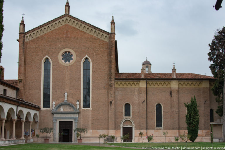 Brick church with two tall pointed windows, central rose window, stone portal, tiled roof, and arcaded walk at left.