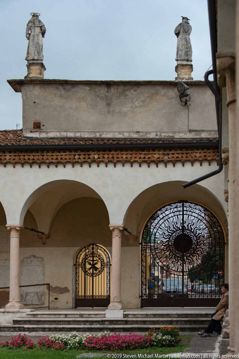 Low arcade with slender columns, two ornate wrought-iron gates, flower beds, and a stone bench.