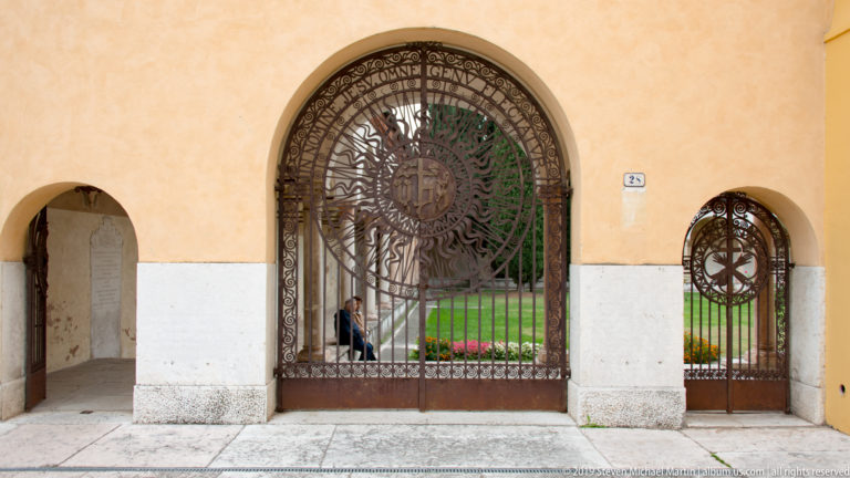 Large and small arched wrought-iron gates set in an ochre wall, both decorated with radiant circular motifs.
