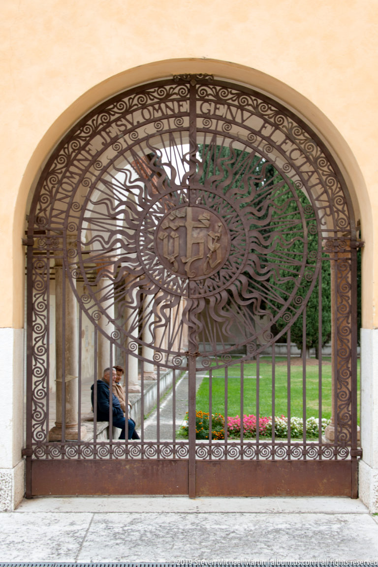 Wide arched wrought-iron gate with dense scrollwork and a large sunburst-like circular emblem at the center.