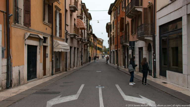 Straight Verona street lined with ochre and brick façades, balconies, awnings, shopfronts, and a few pedestrians.