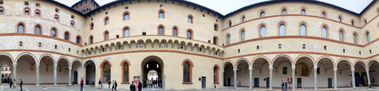 Large cream-colored historic complex with red-trimmed arches, multiple arcades, narrow upper windows, and pedestrians in a courtyard.