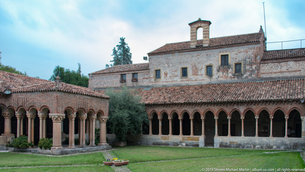Two arcaded sides of a grassy courtyard with stone columns, rounded brick arches, tiled roofs, and taller buildings beyond.