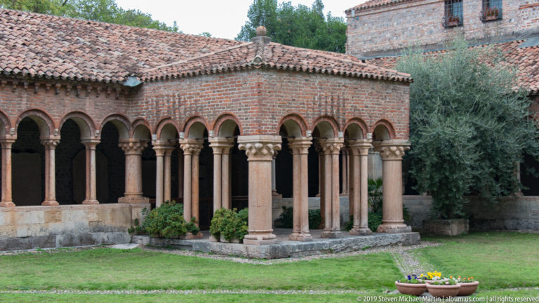 Brick round-arched arcade on stone columns surrounding a lawn with trees and terracotta roofs.