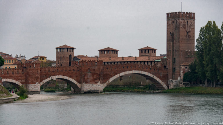 Full view of Ponte Scaligero across the Adige with three arches, crenellated walls, roofed towers, and Castelvecchio keep.