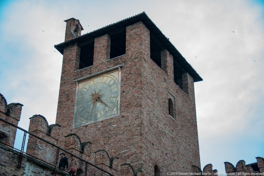 Castelvecchio clock tower with painted dial, open upper belfry, brick walls, and crenellated battlements.