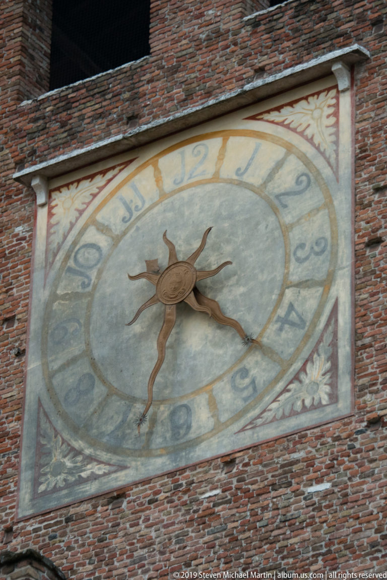 Large painted clock with Roman numerals and sunburst center mounted on a brick Castelvecchio tower.