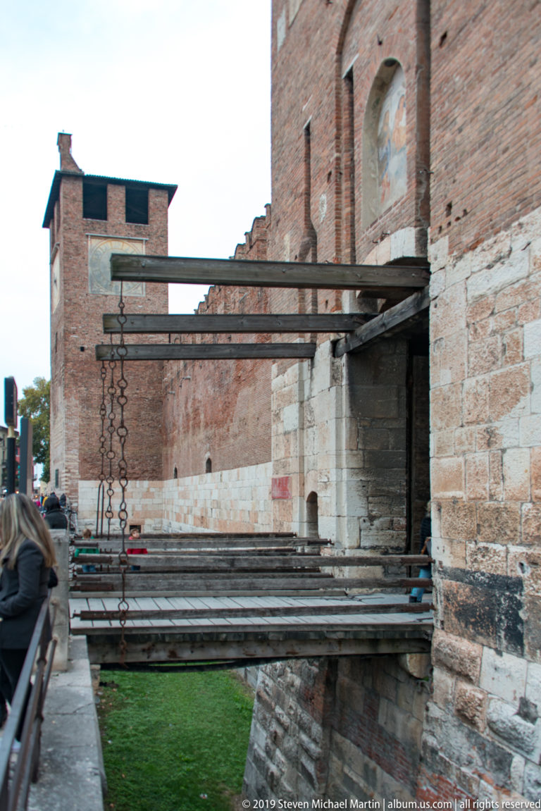 Timber drawbridge with suspension chains crossing Castelvecchio’s moat beside massive brick defensive walls.