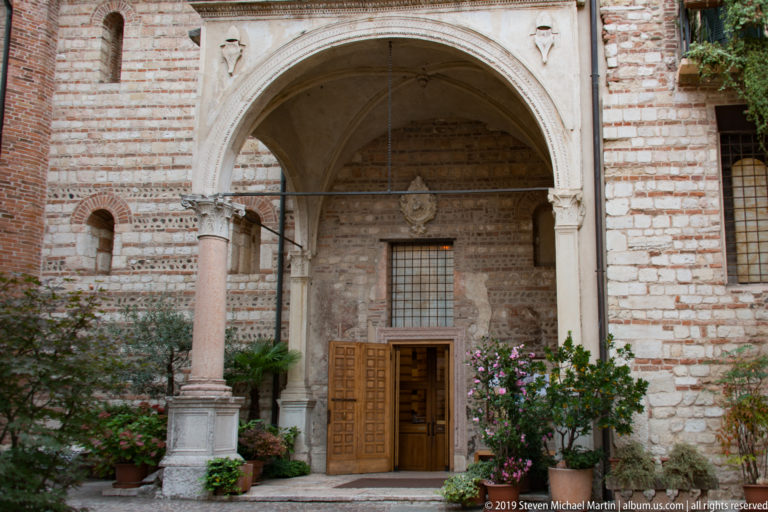 Wooden church doorway beneath a pointed arch, with striped brick-and-stone masonry, columns, and flowering plants.