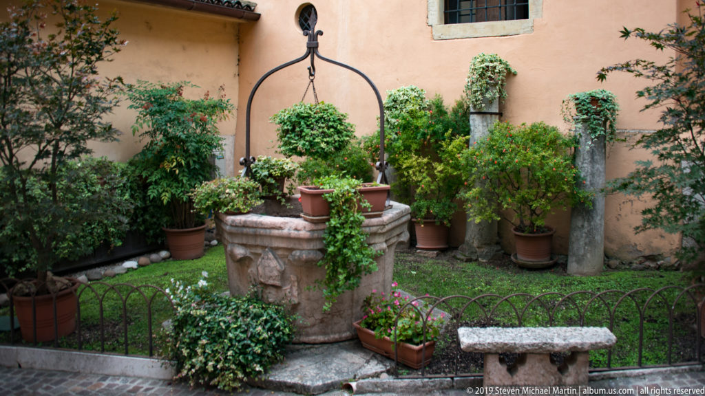 Carved stone wellhead with tall iron support surrounded by potted plants and flowers in a small courtyard.