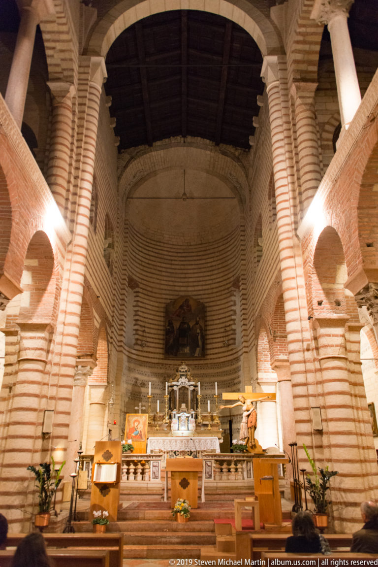 Axial nave view inside San Lorenzo with striped piers, round arches, upper galleries, timber roof, and altar beyond.