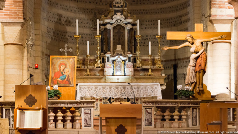 San Lorenzo sanctuary with marble altar, candles, devotional icon, Crucifixion figures, and striped Romanesque masonry.