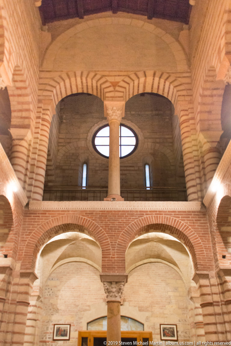 Upper interior of San Lorenzo with striped brick-and-stone arches, central column, gallery opening, and timber roof.