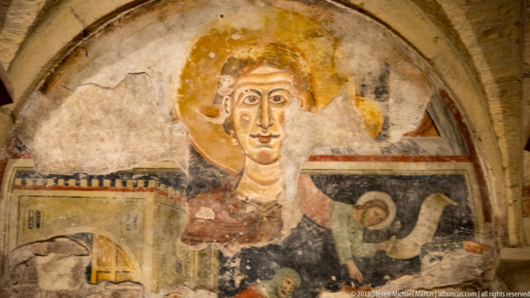 Fragmentary medieval fresco with a large haloed frontal figure surrounded by smaller painted figures and architectural details.