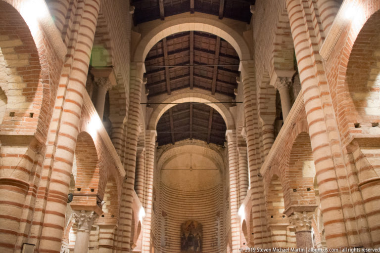 Interior of San Zeno Maggiore with striped brick-and-stone piers, round arches, timber roof, and raised eastern end.
