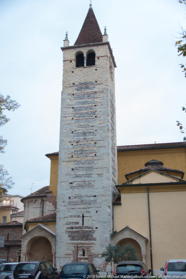 Square medieval bell tower with mixed stone and brick masonry beside a plain ochre church wall in Verona.
