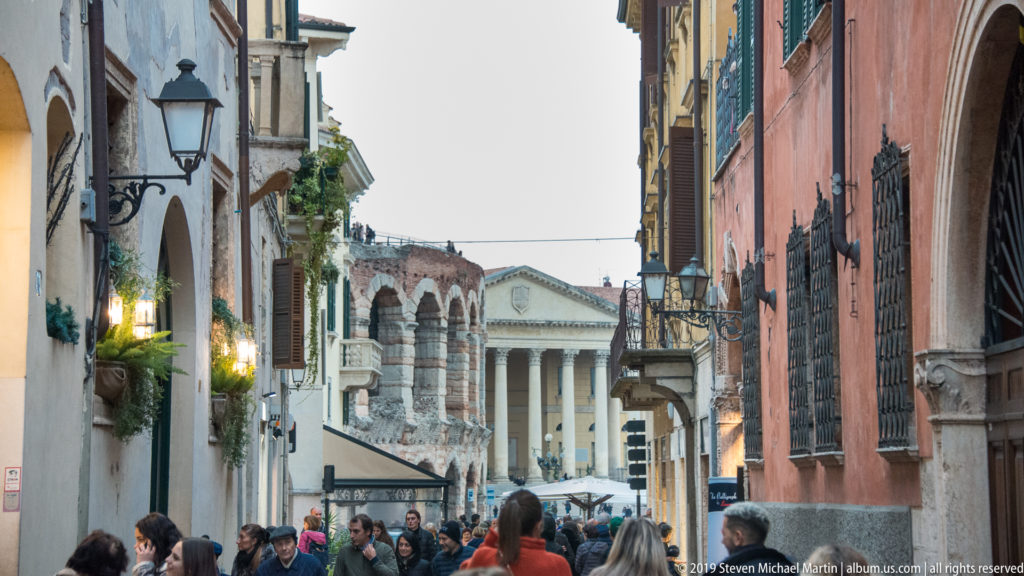 Narrow crowded street opening toward the Arena di Verona and the columned façade of Palazzo Barbieri in Piazza Bra.
