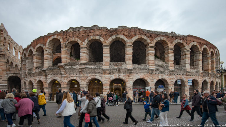 Close exterior view of Arena di Verona with two tiers of weathered arches, entrance signs, and crowds at ground level.