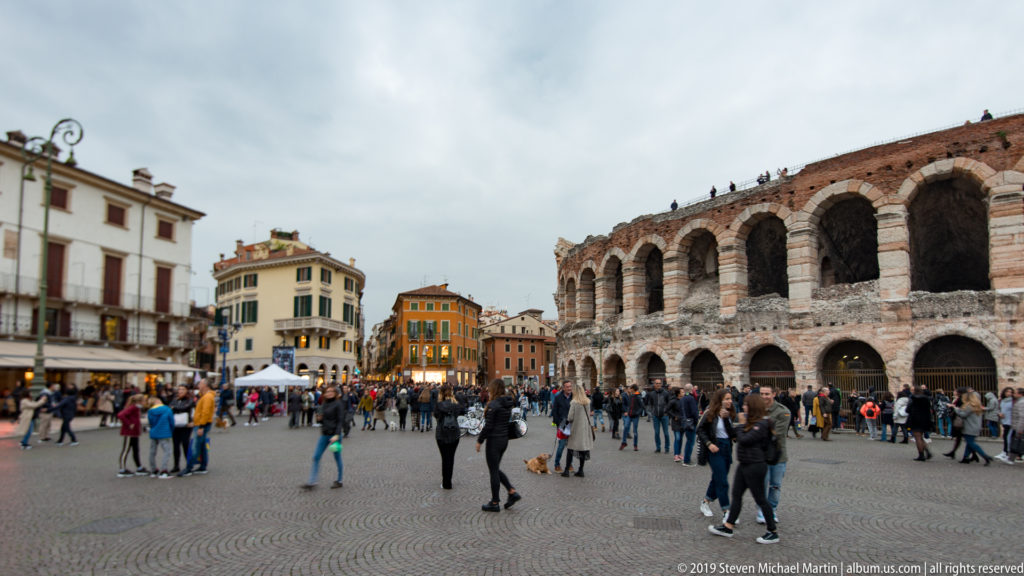 Arena di Verona curving along crowded Piazza Bra with surrounding colorful buildings, stalls, and pedestrians.