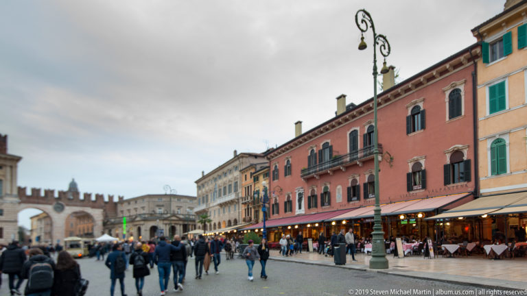 Long view across busy Piazza Bra toward the twin arches of the Portoni della Brà, with cafés and pedestrians at right.