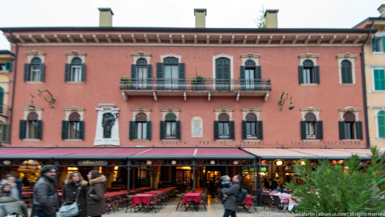 Rose-colored historic façade on Piazza Bra with shuttered windows, balconies, restaurant awnings, and outdoor tables.