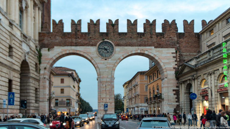 Portoni della Brà with twin stone-lined arches, crenellated brick parapet, central clock, cars, and pedestrians.