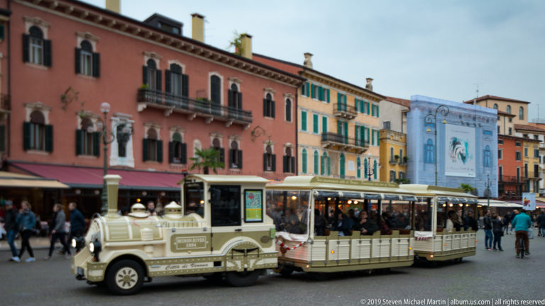Cream sightseeing road train crossing Piazza Bra before colorful façades, cafés, balconies, and crowds of pedestrians.