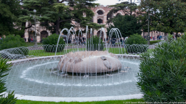 Circular Piazza Bra fountain with arcing jets and the upper arches of the Arena di Verona visible through trees behind.
