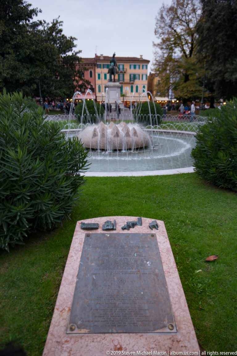 Memorial plaque in grass facing a circular fountain with arcing water jets in the gardens of Piazza Bra.