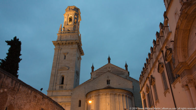 Wide dusk view of Verona Cathedral’s bell tower and apse framed by a dark tree, wall, and neighboring building façades.