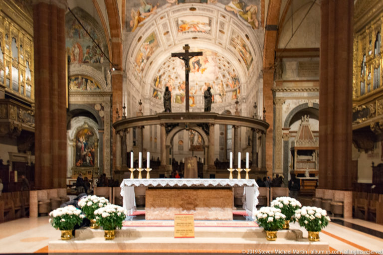 Main altar with white flowers and candles, backed by a circular sanctuary rail, tall crucifix, and frescoed apse.