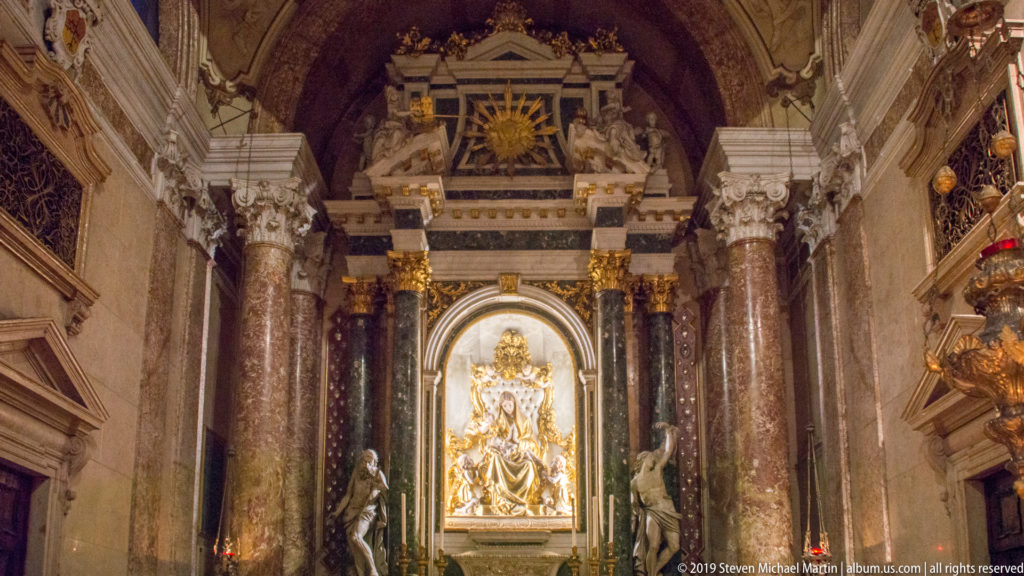 Frontal view of a side altar with tall veined marble columns, bright central sculptural panel, flanking statues, and gilded ornament above.