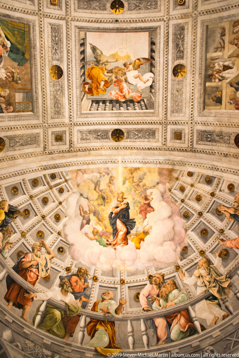 Upward view of a richly painted cathedral apse, with a central figure in clouds, surrounding sacred figures, narrative panels, and decorative coffering.