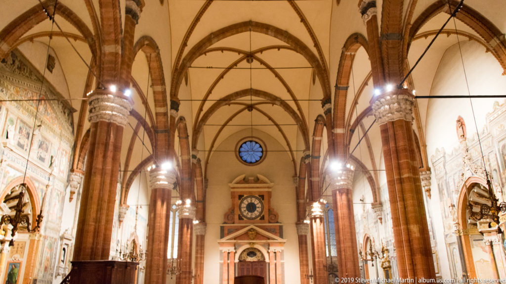 Centered view down Verona Cathedral’s interior with tall red-brick piers, pointed ribbed vaults, side chapels, and a clock at the far end.