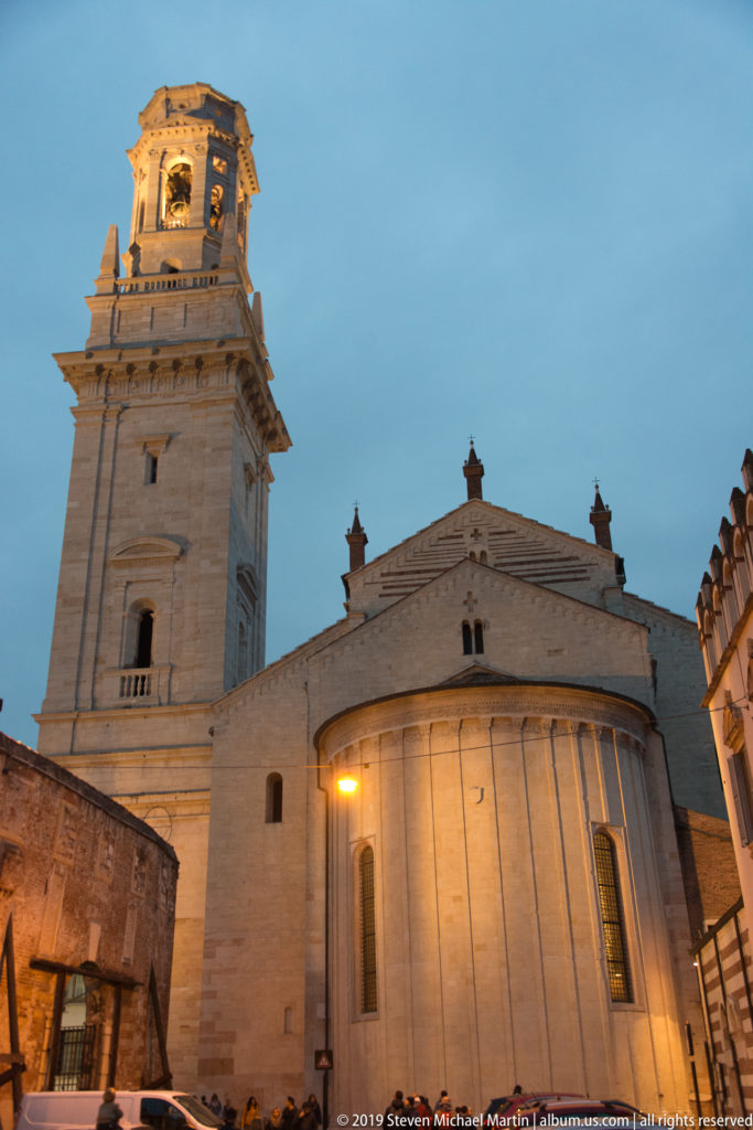 Verona Cathedral’s pale apse and tall square bell tower seen from below at dusk, with warm lights against a blue sky.