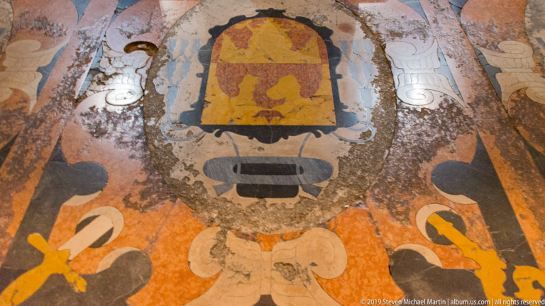 Worn polychrome stone floor inlay with a heraldic shield, ecclesiastical insignia, cords, tassels, and scrolling ornament.
