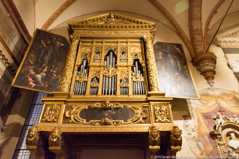 Gilded cathedral pipe organ with vertical metal pipes, carved ornament, sculpted heads, and large paintings on either side.