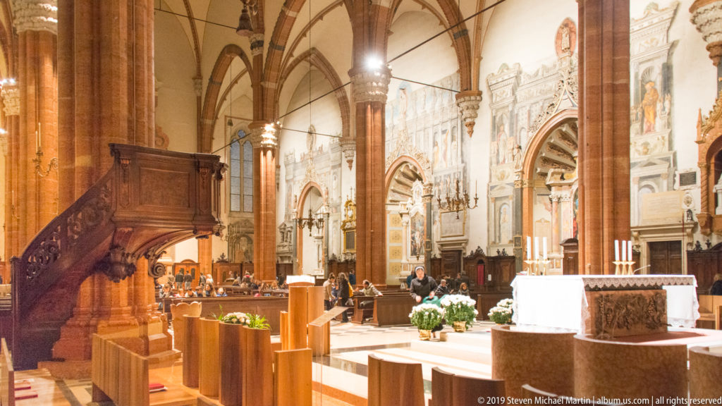 Interior of Verona Cathedral with a large carved wooden pulpit beside red-brick piers, pointed vaults, side chapels, and visitors.