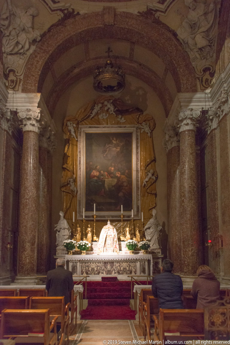 Frontal view of a cathedral altar with large reddish marble columns, central painting, candles, flowers, and seated people in the foreground.