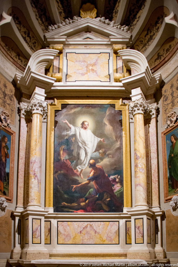Large religious painting framed by paired marble columns and architectural ornament, with smaller paintings on both sides.