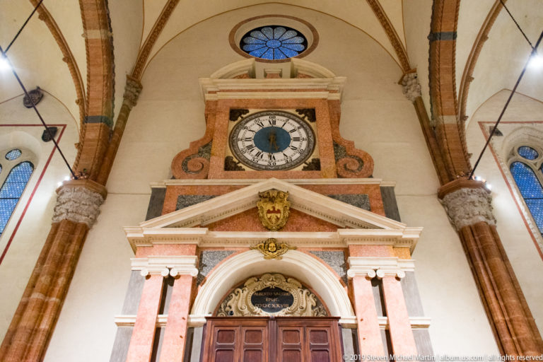 Cathedral wall with a round clock above a pedimented doorway, framed by brick piers and ribbed vaulting, with a rose window overhead.