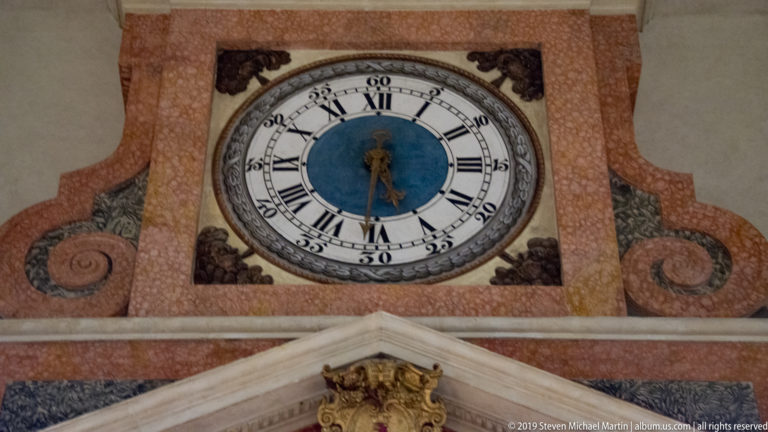 Round cathedral wall clock with Roman numerals and blue center, set within a painted pink-and-gray ornamental surround.