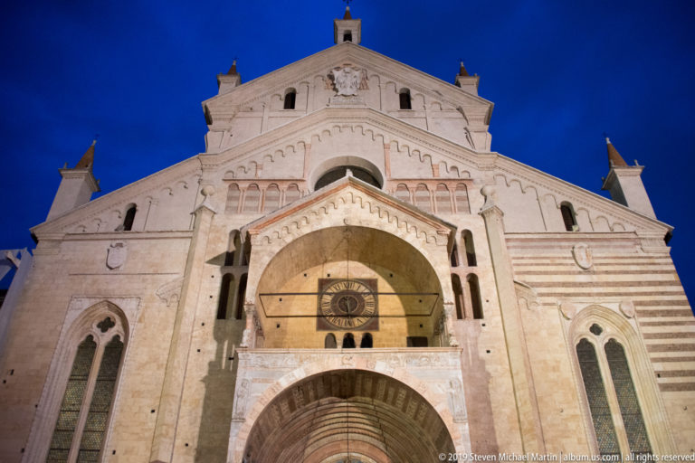 Upper portion of Verona Cathedral’s west façade seen from below, with gable, arcades, narrow openings, and the entrance porch below.
