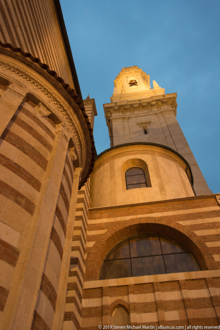 Low-angle exterior view with striped brick-and-stone masonry, a large arched window, curved wall, and illuminated tower against blue evening sky.