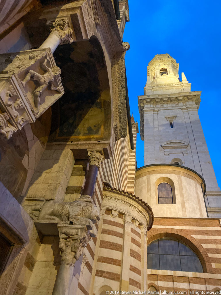 Low-angle exterior view of striped stone-and-brick cathedral masonry beside the tall bell tower under a vivid blue sky.