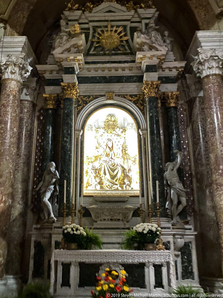 Close view of a marble altar framing a bright sculptural relief, with flanking statues and flower arrangements below.