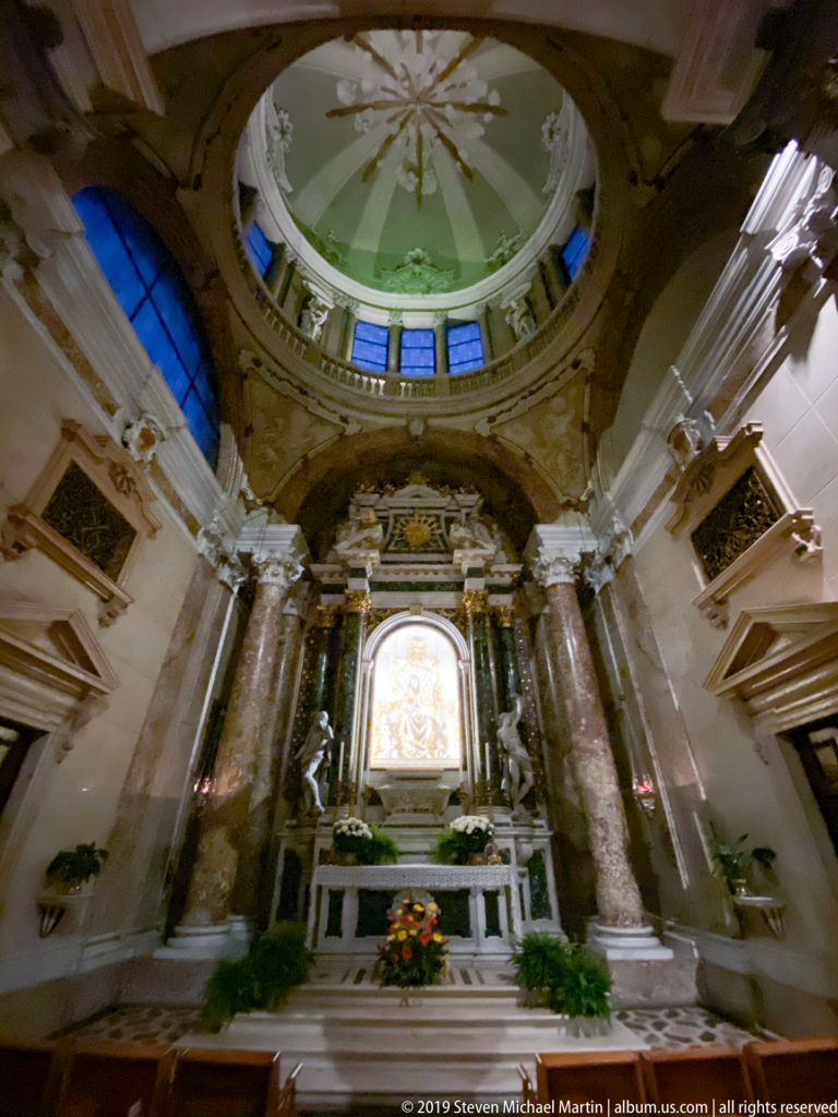 Marble side altar with paired columns, sculptural figures, flowers, and a brightly illuminated central devotional relief beneath a small dome.
