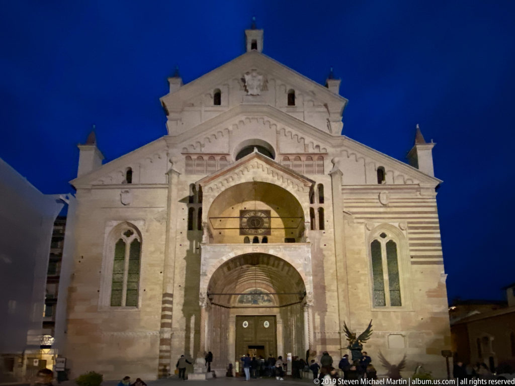 Illuminated pale stone façade of Verona Cathedral beneath a deep blue evening sky, with people gathered near the recessed entrance.
