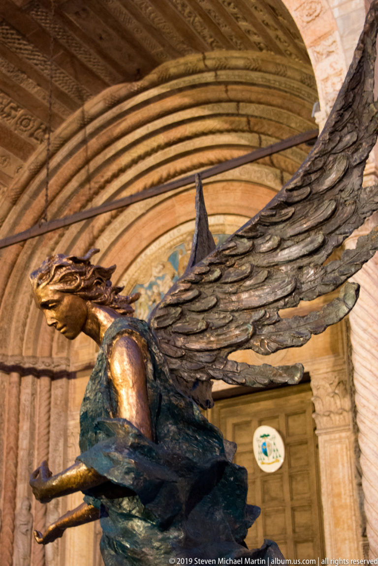 Close low-angle view of a dark bronze winged figure with outstretched arms and large feathered wings beneath a carved stone arch.