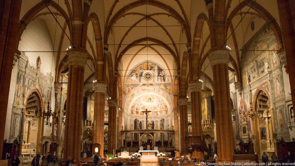 Central nave of Verona Cathedral with tall brick piers and ribbed vaults leading toward the illuminated altar and frescoed apse.